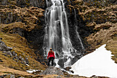 Blick von hinten auf eine Frau, die vor einem Wasserfall an der Strandir-Küste in der Nähe der Stadt Djupavik im Nordwesten von Island steht; Djupavik, Westfjorde, Island