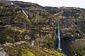 Woman standing and overlooking Mulagljufur Canyon, a hikers paradise, watching an amazing view of a waterfall and the moss-covered cliffs; Vik, South Iceland, Iceland