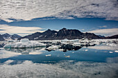 Jagged mountain peaks and white clouds reflected in the the calm waters in Nansen Fjord with icebergs and growlers floating in the foreground at the point where the fjord's glacier enters the water; East Greenland, Greenland