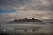 Jagged mountain peaks and foggy clouds reflected in the calm waters along Nansen Fjord; East Greenland, Greenland