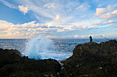 Waves crash against the coast near Folly Lighthouse as a fisherman stands on the rocky shore; Port Antonio, Jamaica