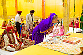 Ein Hochzeitshelfer führt ein traditionelles Ritual bei einer indischen Hochzeitszeremonie durch, während die Braut und der Bräutigam zusehen; Ludhiana, Punjab, Indien