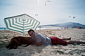 Ein Mann liegt am Strand von Punta Paloma mit seinem Hund unter einem Sonnenschirm; Tarifa Cadiz Andalusien Spanien