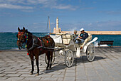 Greece, Crete, Hania, Two men sitting in horse drawn carriage, lighthouse and harbor in background.