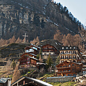 Häuser an der Seite eines Hügels mit einem weißen Kreuz auf einem hohen Felsvorsprung; Zermatt Wallis Schweiz