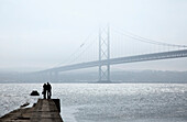 Silhouette eines Paares, das am Ende eines hölzernen Piers steht und über das Wasser auf eine Brücke im Nebel hinausblickt; North Queensferry Fife Schottland