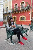 Young Korean woman sitting on park bench in old spanish town looking at cell phone; Guanajuato, Guanajuato State, Mexico