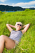 A Young Woman Lays On The Grass With A View Of The Coastline; Urupukapuka Island, New Zealand