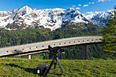 Eine Kamera und ein Stativ auf einem grasbewachsenen Hügel, um die verschneite Bergkette einzufangen; San Gottardo, Tessin, Schweiz