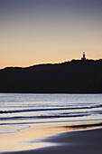 Silhouette des Cape Byron Leuchtturms auf der Bergkuppe und Strand bei Sonnenuntergang in Byron Bay in New South Wales, Australien