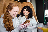 Lächelnde junge Frauen telefonieren und schauen in die sozialen Medien im Cafe