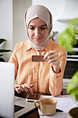 Lächelnde Frau mit Hidschab hält Kreditkarte beim Online-Shopping
