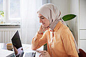 Lächelnde Frau mit Hidschab bei der Heimarbeit mit Laptop