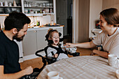Lächelnde Eltern mit glücklichem behindertem Kind im Rollstuhl am Küchentisch sitzend