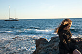 Frau auf Felsen am Meer sitzend mit Winken und Segelboot im Hintergrund