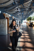 Lächelnde Frau benutzt Handy beim Gehen am Bahnhof