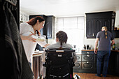 Parents with disabled teenage daughter in wheelchair in kitchen, mother feeding daughter in wheelchair while father preparing food