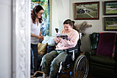 Mutter mit behinderter Tochter im Rollstuhl im Wohnzimmer, jugendliches Mädchen liest Zeitschrift