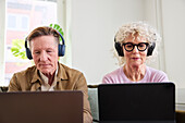 Älterer Mann und Frau sitzen im Wohnzimmer und benutzen Laptop und digitales Tablet, um Podcasts zu bearbeiten