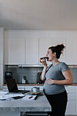 Schwangere Frau arbeitet von zu Hause aus, während sie frühstückt