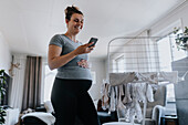 Pregnant woman using phone while doing housework and hanging baby clothes on drying rack