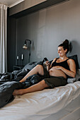 Glückliche schwangere Frau entspannt sich im Bett und benutzt das Telefon
