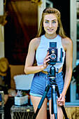 Portrait einer jungen schönen kaukasischen Frau in den 20ern mit blauen Augen, die mit einer alten Vintage-Kamera auf einem Stativ im Freien fotografiert. Lifestyle-Konzept.