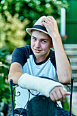 Portrait of young caucasian boy with a broken and cast arm wearing a hat and sitting in a chair outdoor in a garden. Lifestyle concept.