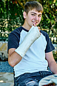 Porträt eines jungen kaukasischen Jungen mit gebrochenem und eingegipstem Arm, der auf einem Stuhl im Freien in einem Garten sitzt. Lebensstil-Konzept.