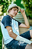 Porträt eines jungen kaukasischen Jungen mit gebrochenem und eingegipstem Arm, der einen Hut trägt und auf einem Stuhl im Freien in einem Garten sitzt. Lebensstil-Konzept.