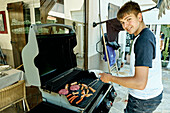 Porträt eines kaukasischen Jungen beim Grillen von Fleisch im Garten eines Landhauses. Lifestyle-Konzept.