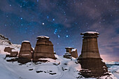 Orion rising behind the iconic Hoodoos on Highway 10 east of Drumheller, Alberta, near East Coulee, on a moonless January night, with illumination by starlight and by a nearby yardlight providing some shadows and warmer illumination. Clouds are beginning to move in and are providing the natural star glows.
