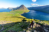 Wanderer steht auf dem Gipfel eines Hügels mit dem Dorf Vidareidi und dem Berg Malinsfjall im Hintergrund, Insel Vidoy, Färöer, Dänemark, Europa
