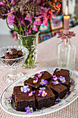 Brownies garniert mit essbaren Blüten auf Porzellanplatte, dekorative Kerze im Hintergrund