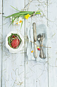 Easter nest in paper bag with red Easter egg, bouquet of daffodils and cutlery on linen napkin