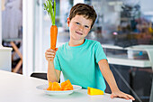 Boy in casual clothes smiling and looking at camera while standing at kitchen table with grater and sliced carrot in plate