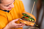 Crop positive young female in casual clothes with delicious burger in cafe