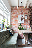 Dining area with brick wall, wooden table and green upholstered bench, plants on windowsill