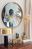 Round mirror with golden frame above decorated sideboard with dried flowers and golden table lamp