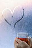 Close up hands cupping hot apple cider below heart shape on wet window\n