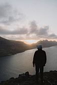 Mann auf dem Gipfel eines Hügels mit Blick auf den Sonnenuntergang über dem Fluss, Klakkur, Klaksvik, Färöer Inseln