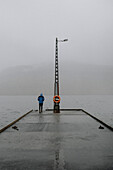 Mann steht im Regen auf einem Steg mit Blick auf den nebligen Ozean, Kollafjorour, Färöer Inseln