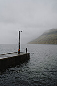 Man standing on rainy, foggy jetty over ocean, Kollafjorour, Faroe Islands\n