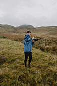Männlicher Fotograf mit Spiegelreflexkamera auf grasbewachsenem Hügel, Durness, Schottische Highlands, Schottland