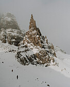 Hikers climbing mountain slope below snow covered rock formation, Old Man of Storr, Scotland\n