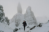 Male photographer hiking below snow covered rock formation in mountains, Old Man of Storr, Scotland\n