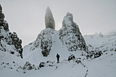 Hiker standing below tall, snow covered mountain rock formation, Old Man of Storr, Scotland\n