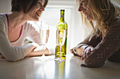 Zwei junge Frauen sitzen am Tisch, unterhalten sich lachend und trinken Wein