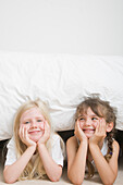 Two young girls with chin resting on hands under bed smiling\n