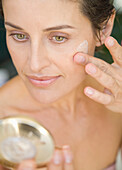 Close up of woman applying moisturizing cream and looking into mirror\n